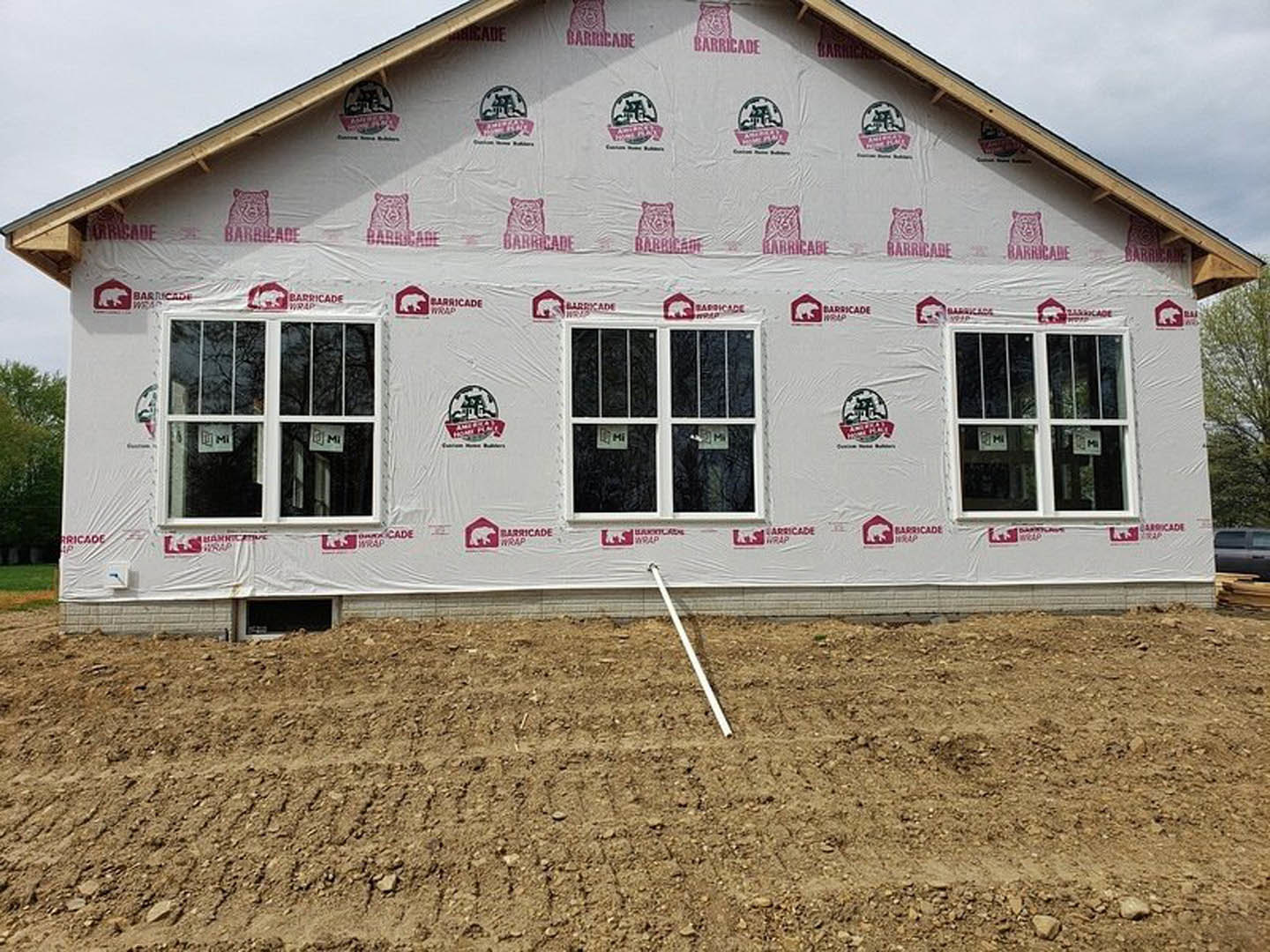 White-sided house with large windows, patch of bare dirt in foreground, white pole standing upright, small sign taped to window, black truck partially visible, trees and grass