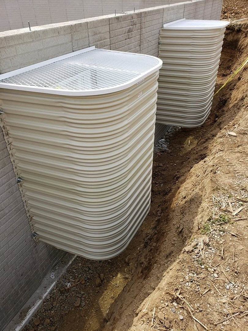 White pipes mounted on a brick wall beside a white plastic tank with a metal grate, set on a concrete outdoor surface.