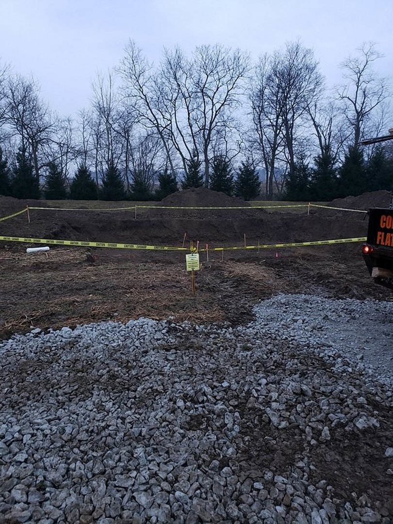 Gravel and dirt construction site bordered by yellow caution tape, surrounded by tall trees and forested landscape, under a clear sky