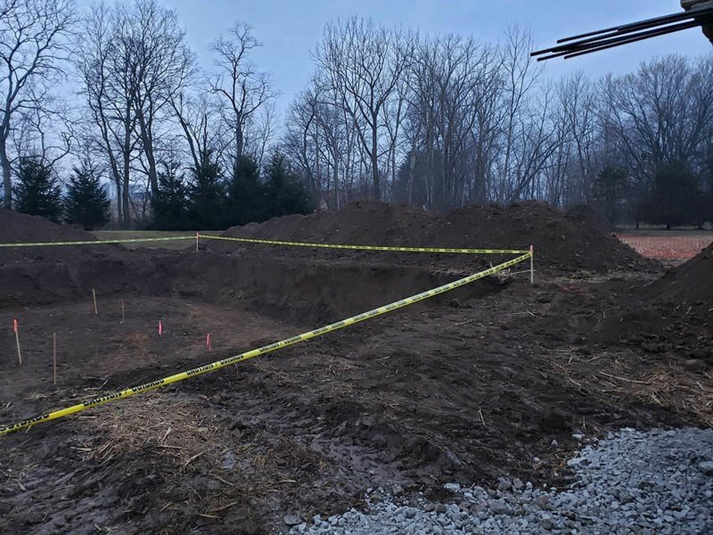 Yellow caution tape stretched across a dirt pit surrounded by bare soil, scattered rocks, and trees in the background under a cloudy sky
