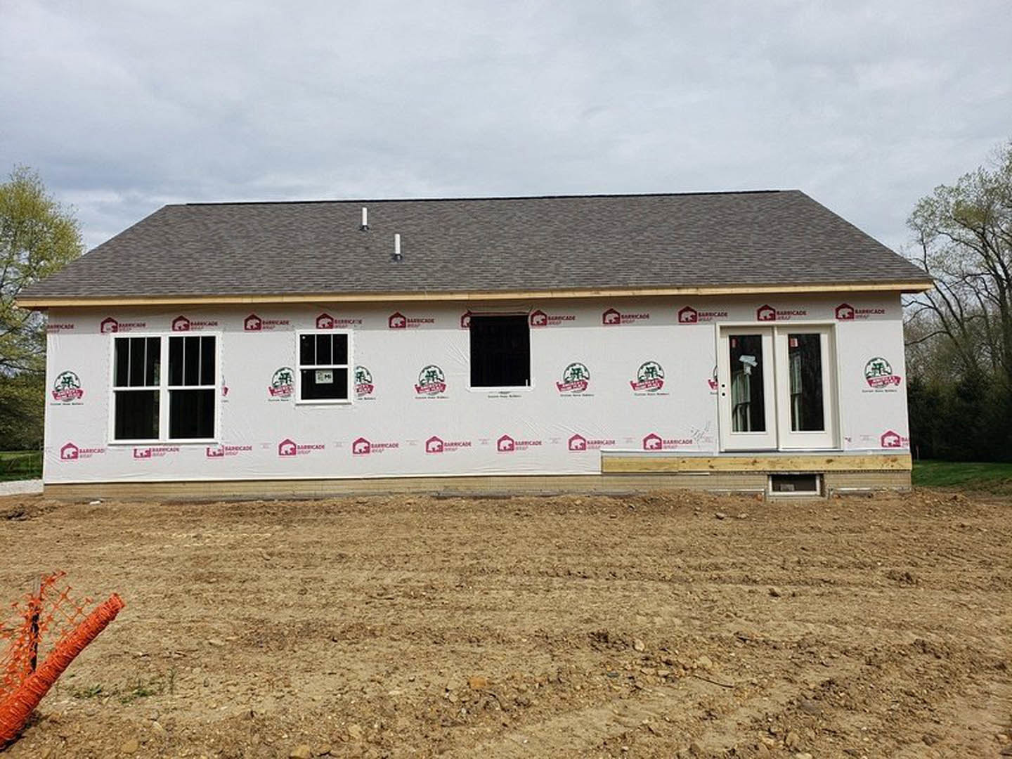 Partially built house with white exterior walls, exposed roof framing, and dirt field in foreground marked by tire tracks, under cloudy sky.