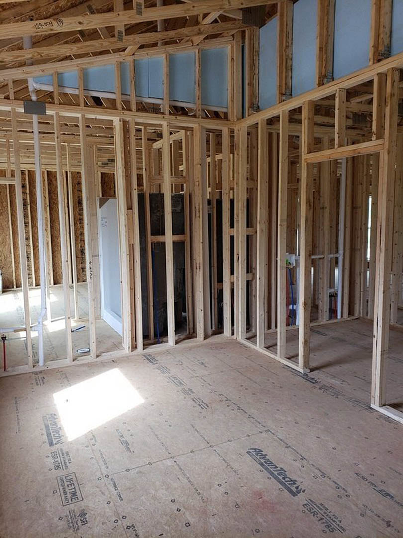 Wood-framed room under construction with exposed beams, unfinished floor, white and blue walls, window close-up, and white pipe visible.