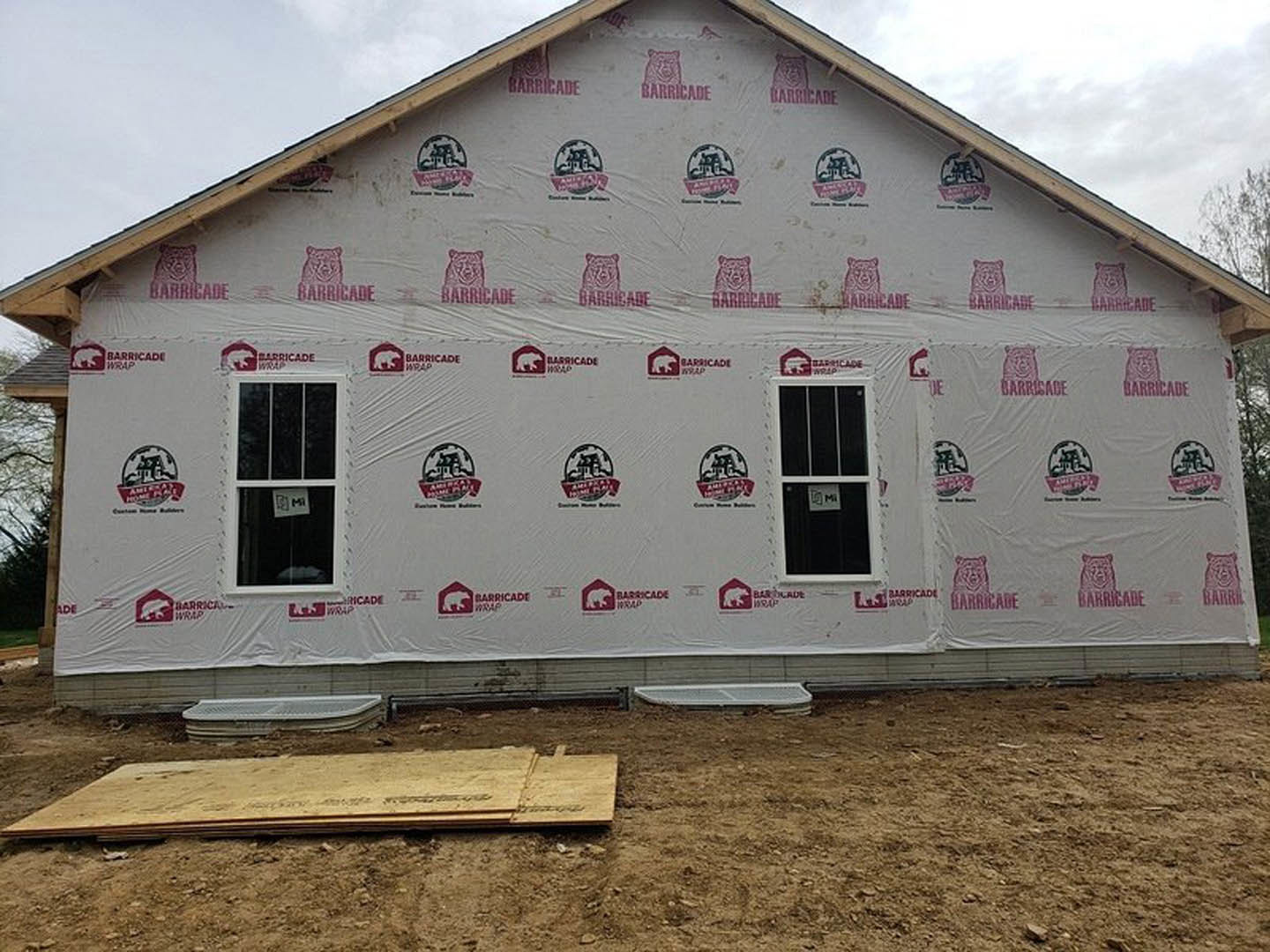 Two-story house wrapped in white plastic sheeting, exposed plywood near window with white sign, pile of lumber on ground, cloudy sky and trees in background.