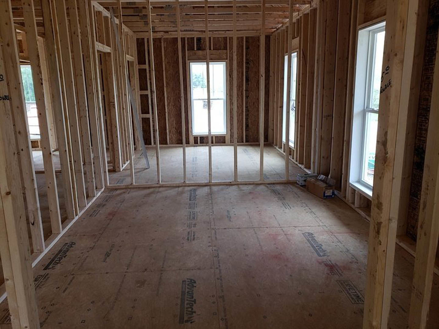Wood-framed room with exposed beams, white metal bars across the floor, large window with glass pane and white frame, wooden crate in foreground, daylight illuminating unfinished
