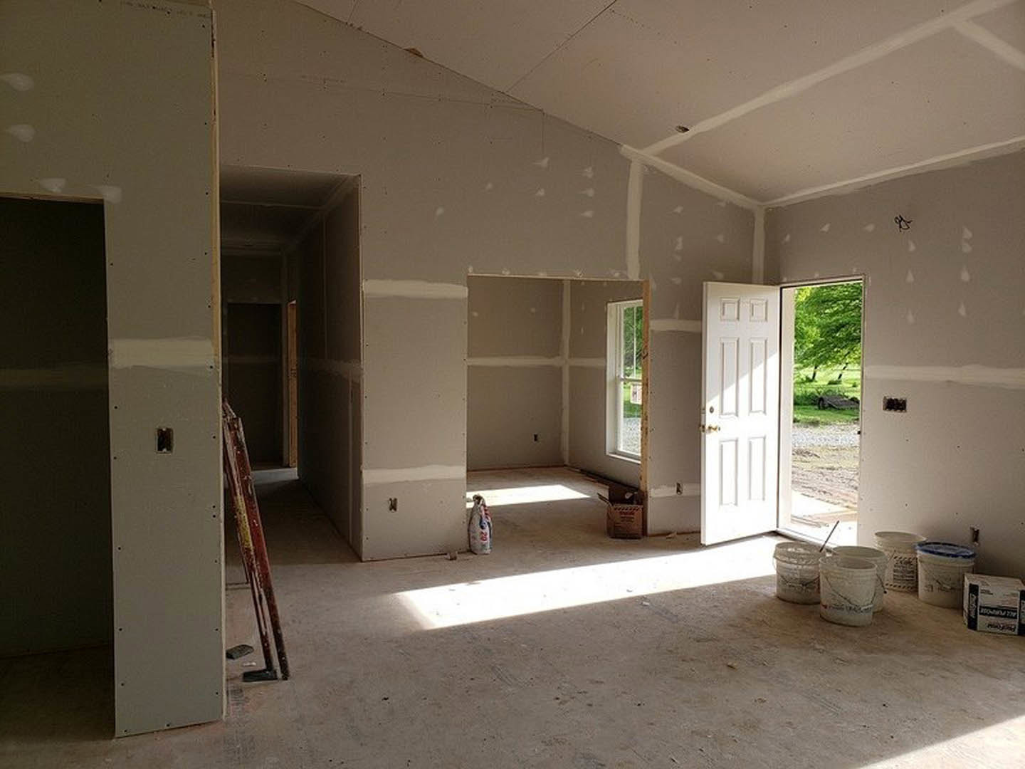 Unfinished interior with painted drywall, scattered paint buckets, white door, red and white pole, and construction materials on bare flooring
