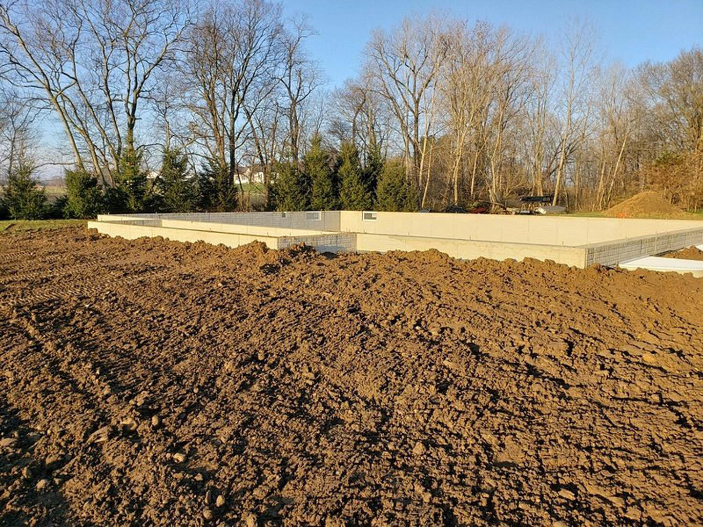 Dirt field with large soil pile, bare trees in background, white rectangular object on ground, sparse grass and natural landscape