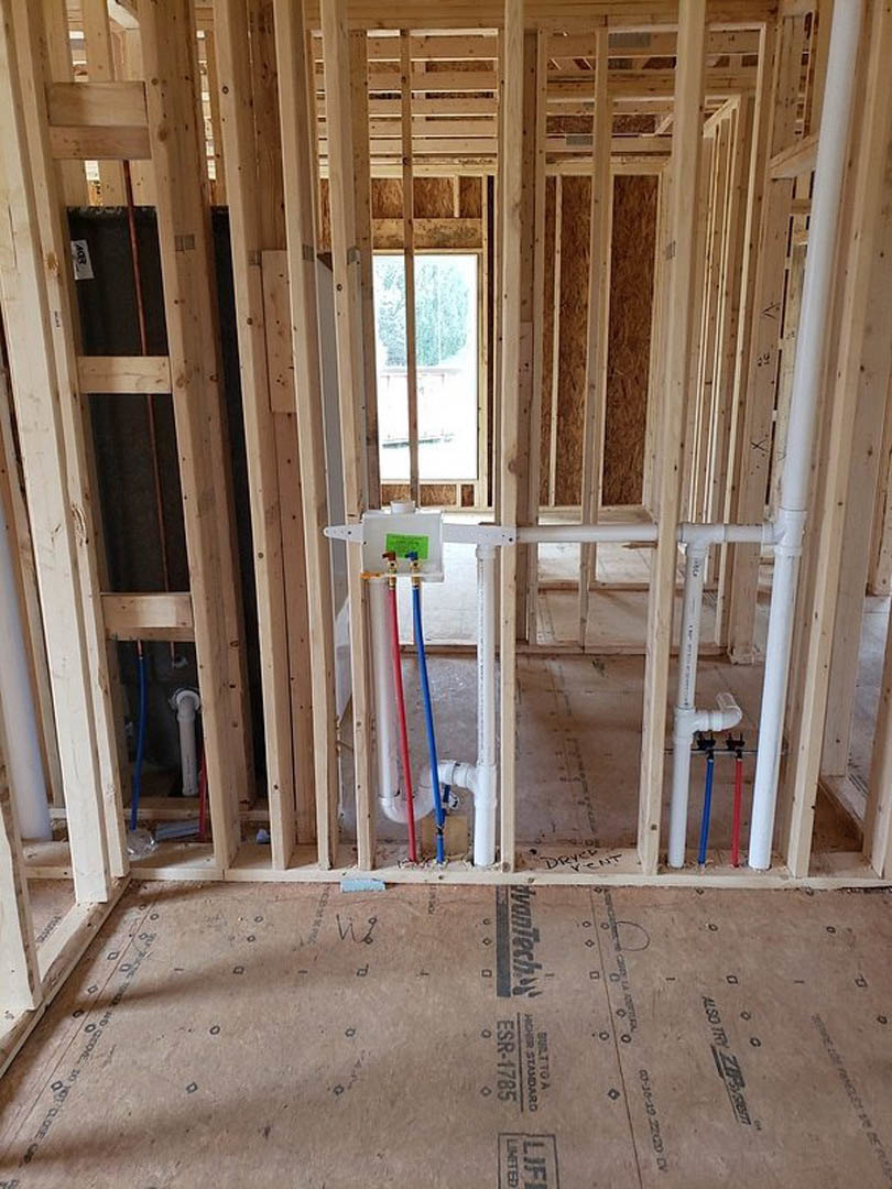 Unfinished interior with exposed wooden beams, construction insulation, visible plumbing pipes, scattered cleaning tools, and a cardboard box on subfloor.