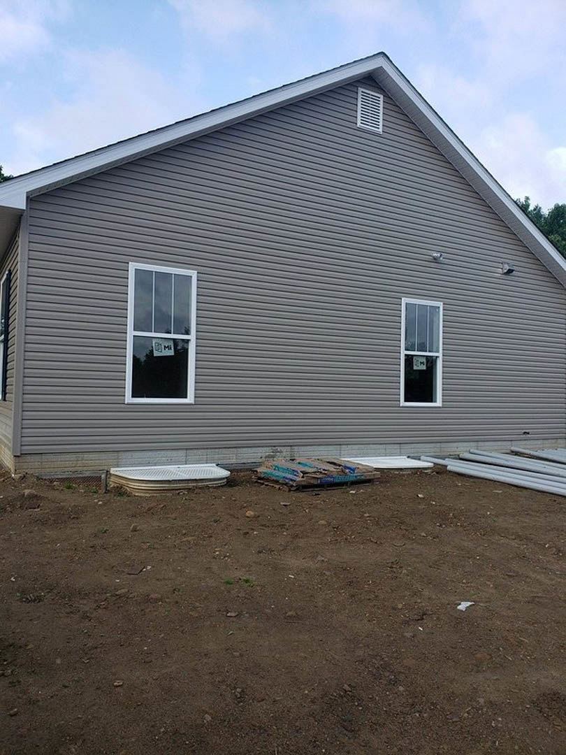 Modern home exterior with minimal windows, gray siding, metal grate detail, dirt yard scattered with pallets and a white tub, cloudy sky overhead