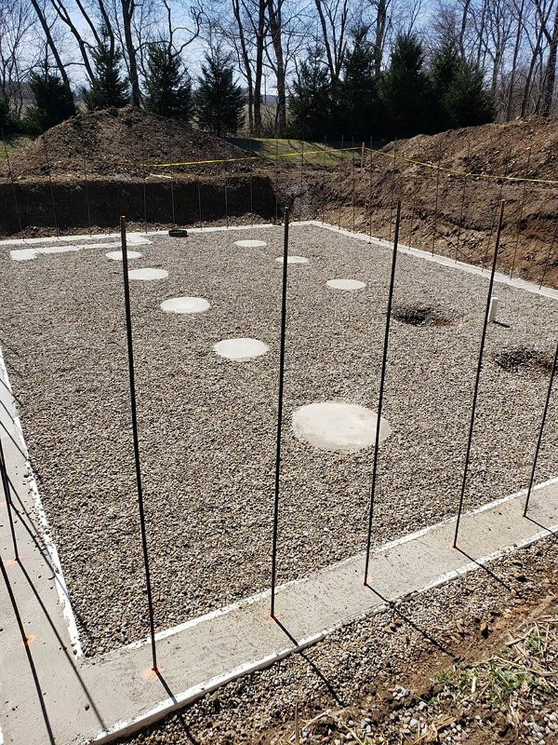 Concrete foundation with metal poles surrounded by piles of gravel, forest trees in background, white marking on ground, close-up of sidewalk and concrete curb