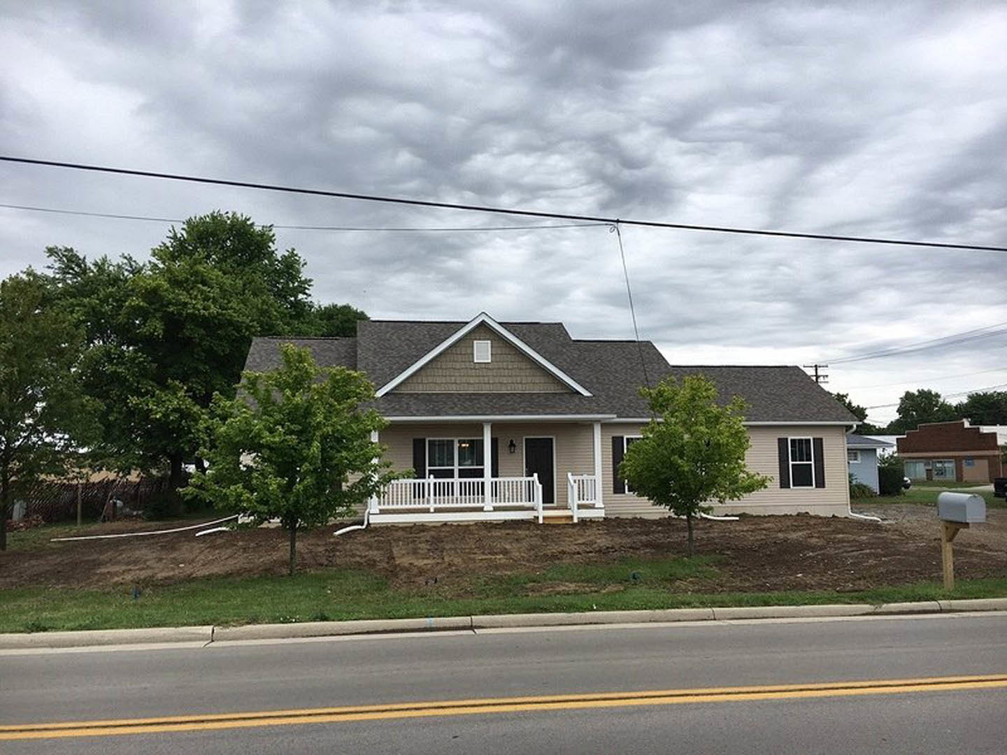 White porch with white railing, gray siding, large front windows, tree in landscaped yard, yellow double line on road in foreground