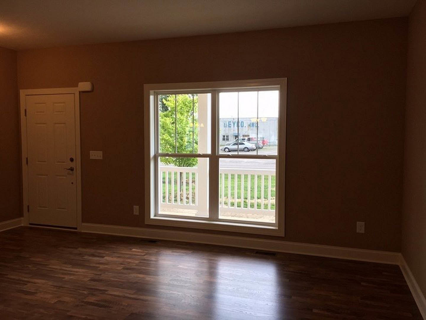 Sunlit room featuring wood flooring, white door with silver handle, large window with white railing, and view of green tree and parked car outside.