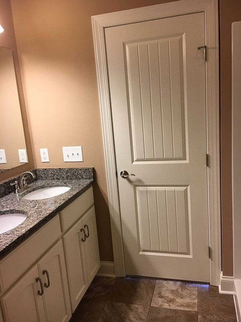 White bathroom with brown tile floor, white wall, close-up of rectangular sink and chrome faucet, white door with silver handle, white electrical outlet with black spots.