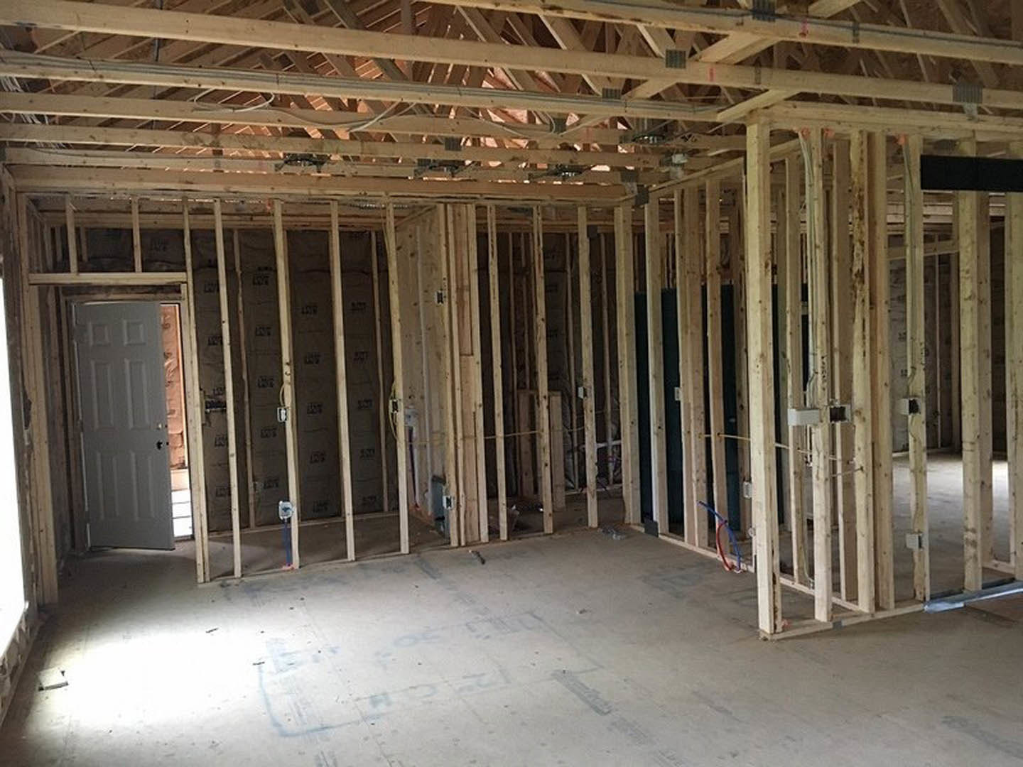 Wood-framed room under construction with exposed beams, white door featuring black handle, concrete floor, and visible insulation.