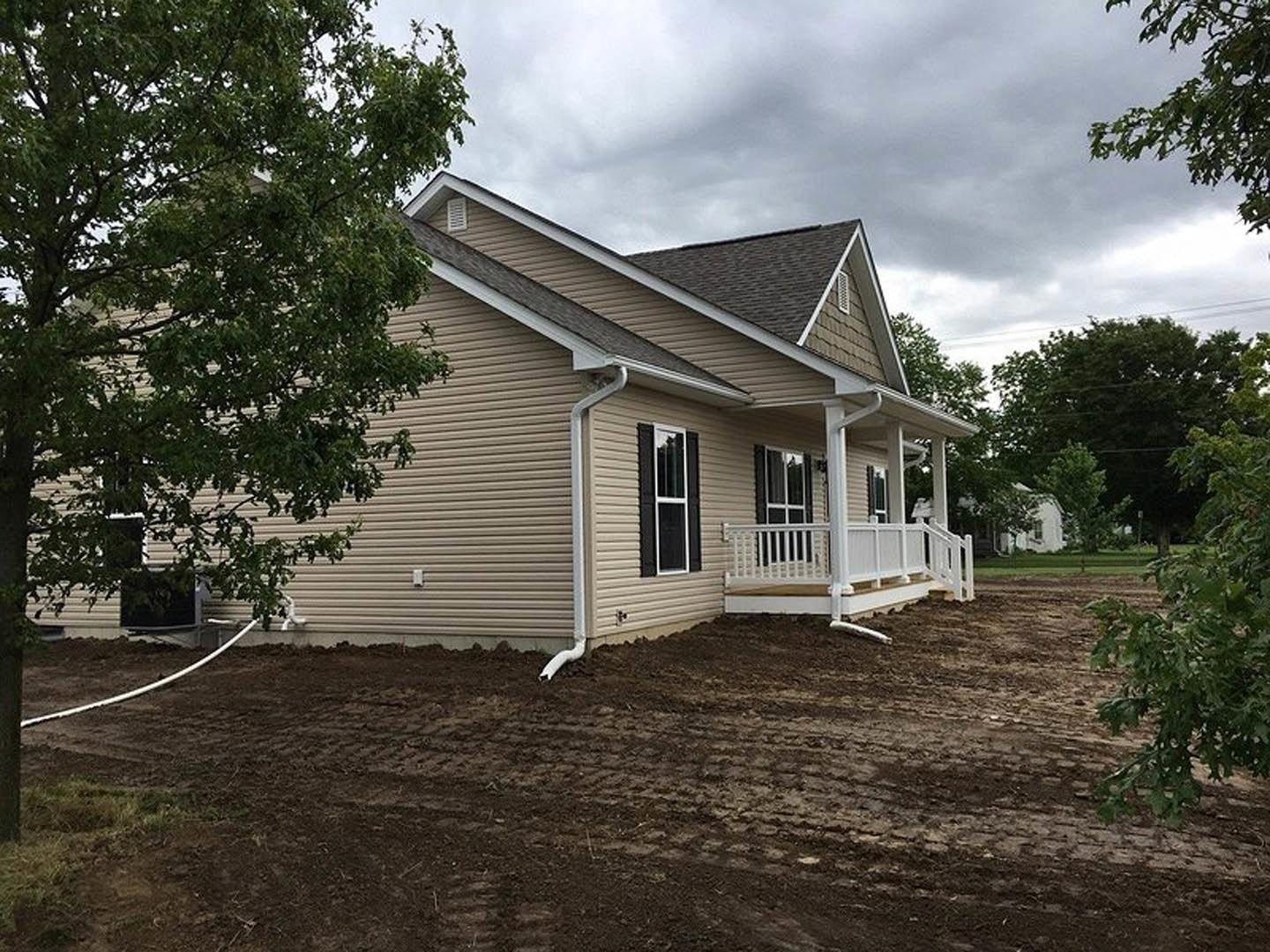 White porch with white railing, large front window, white gutters, dirt patch and tree in front yard, cloudy sky above