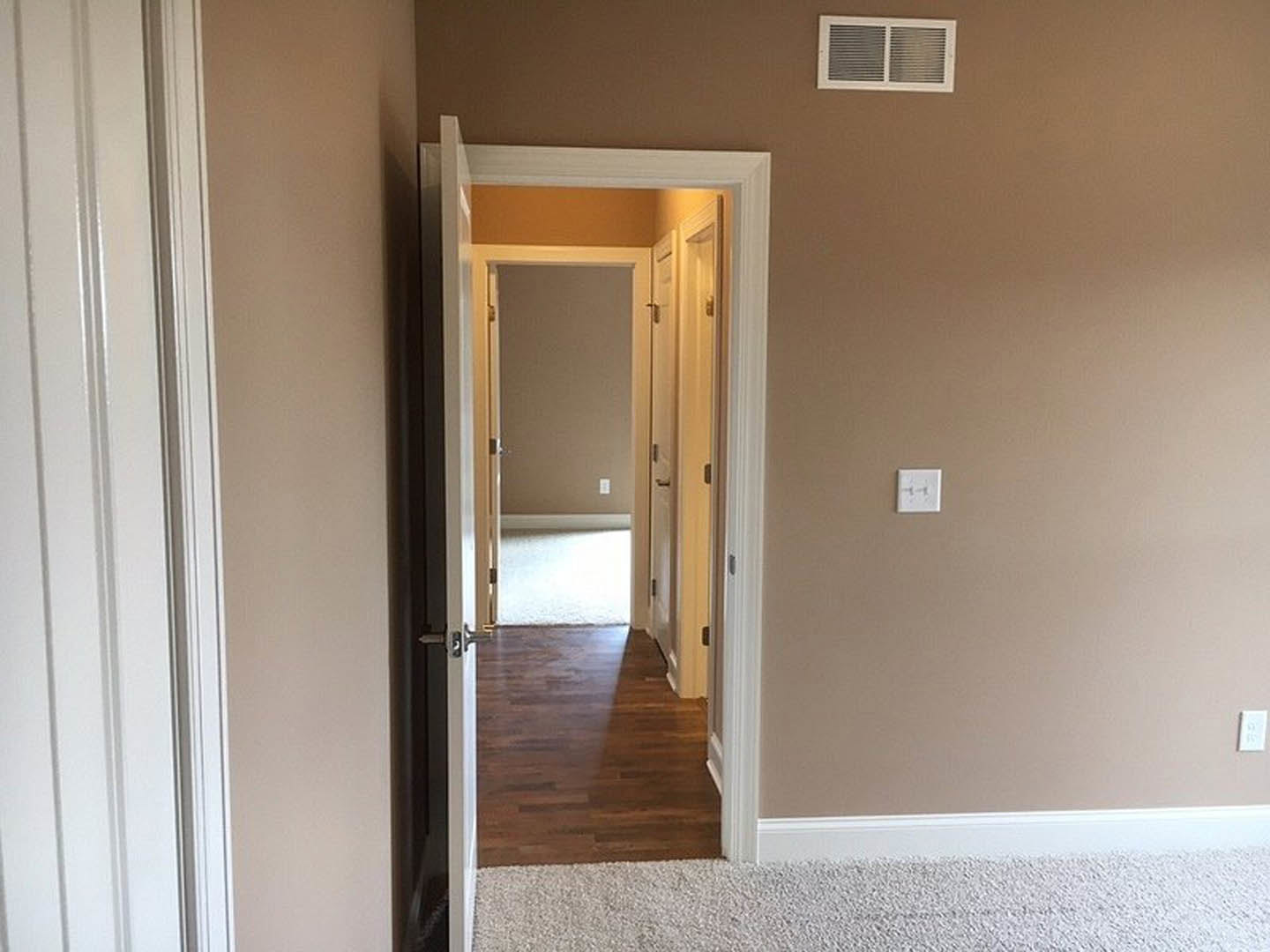 Open white door with black hardware in hallway, wood flooring with sunlight, white walls, ceiling vent, and adjacent white carpet