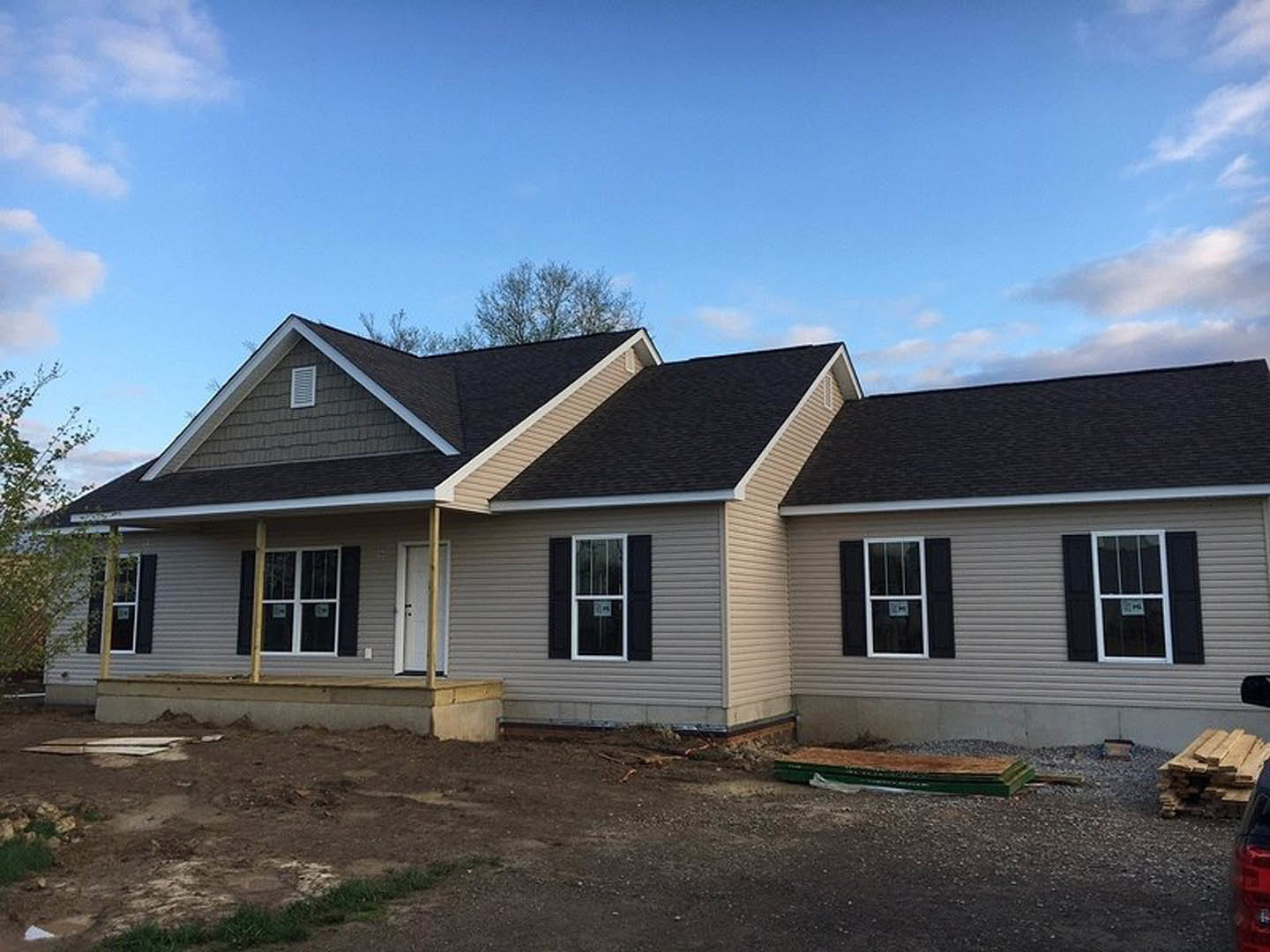 Partially built house with black shingle roof, white-framed windows, and exposed siding, situated beside a dirt road under a cloudy sky