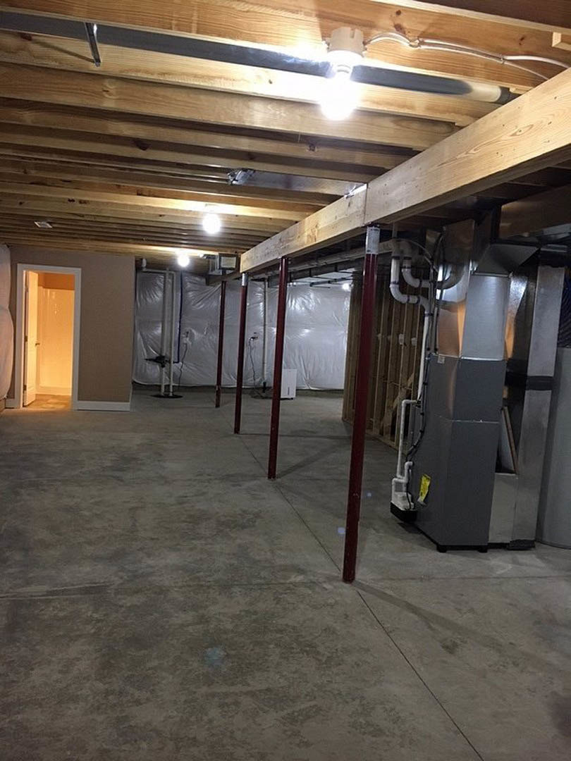 Basement with exposed wood beam ceiling, white plastic wall covering, concrete floor, red steel support poles, and a door leading to an adjacent room