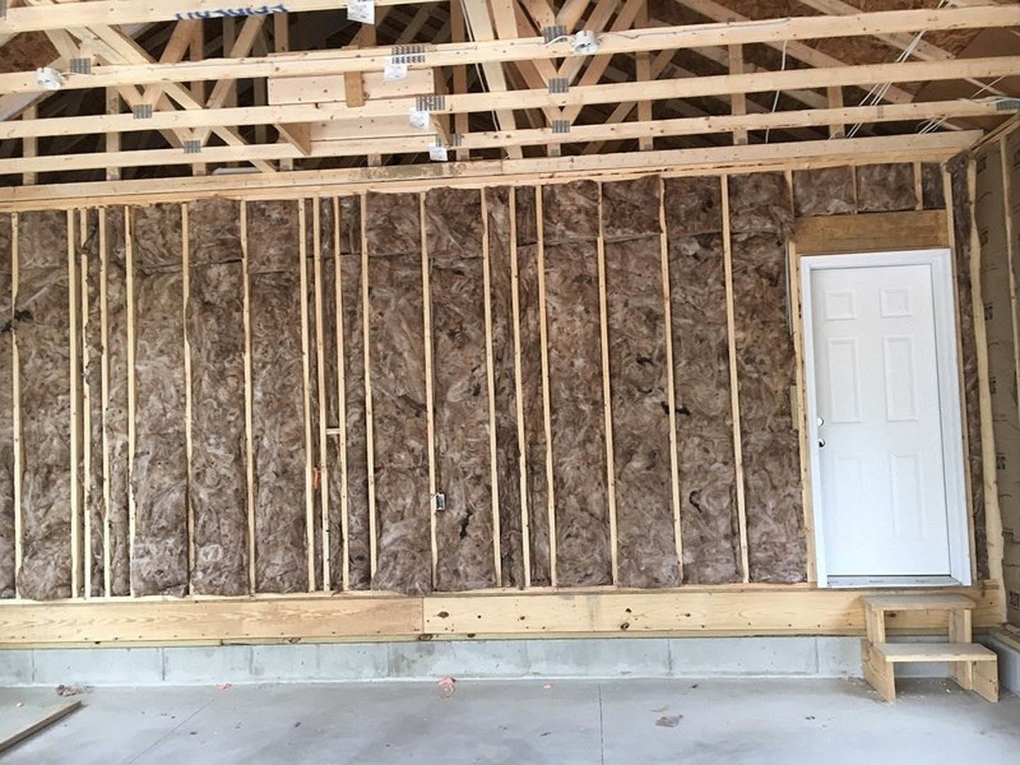 Wood-framed wall with exposed insulation, white door with metal handle, wooden roof beams, concrete floor, and wooden step stool in corner
