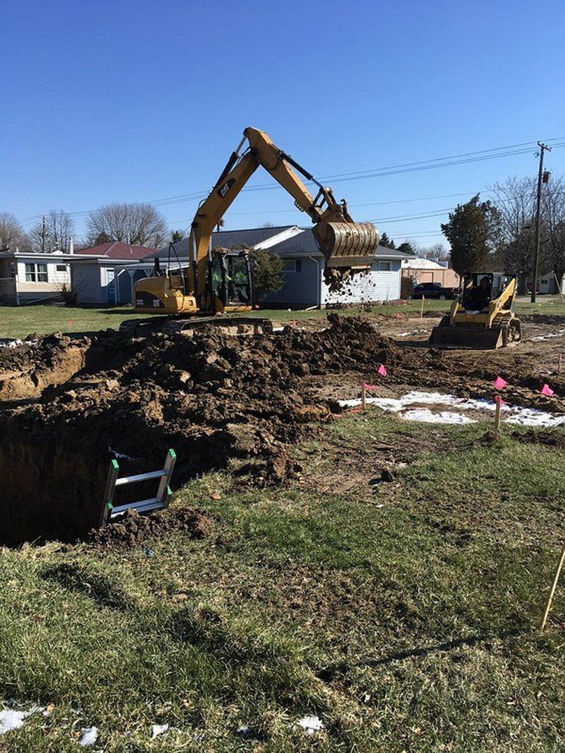 Bulldozer excavating soil on residential construction site, ladder resting in dirt, grassy field and trees under clear blue sky