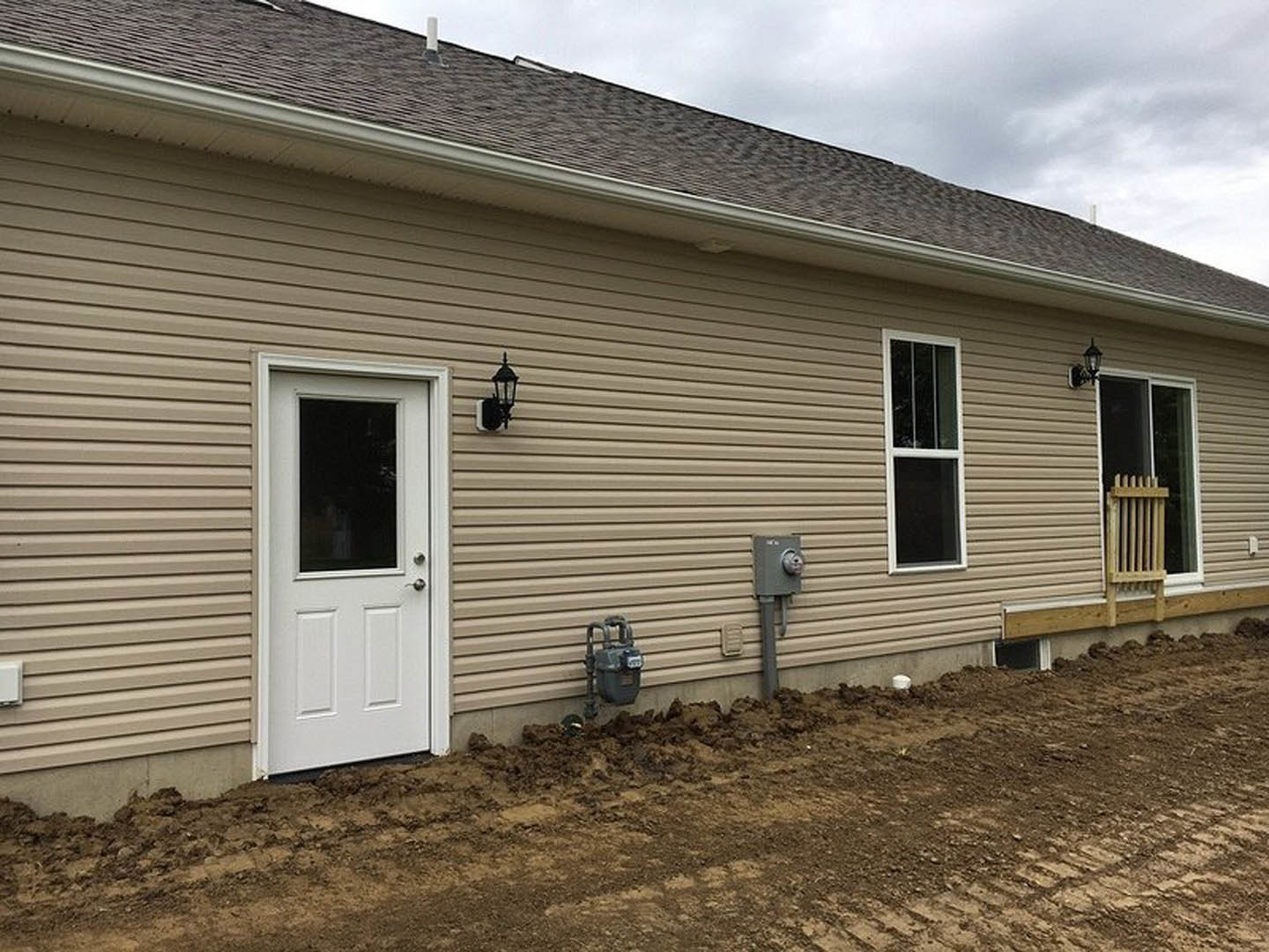 Two-story home with light siding, white framed windows, and a white entry door facing a dirt driveway; grey metal utility box and water meter mounted on exterior wall.