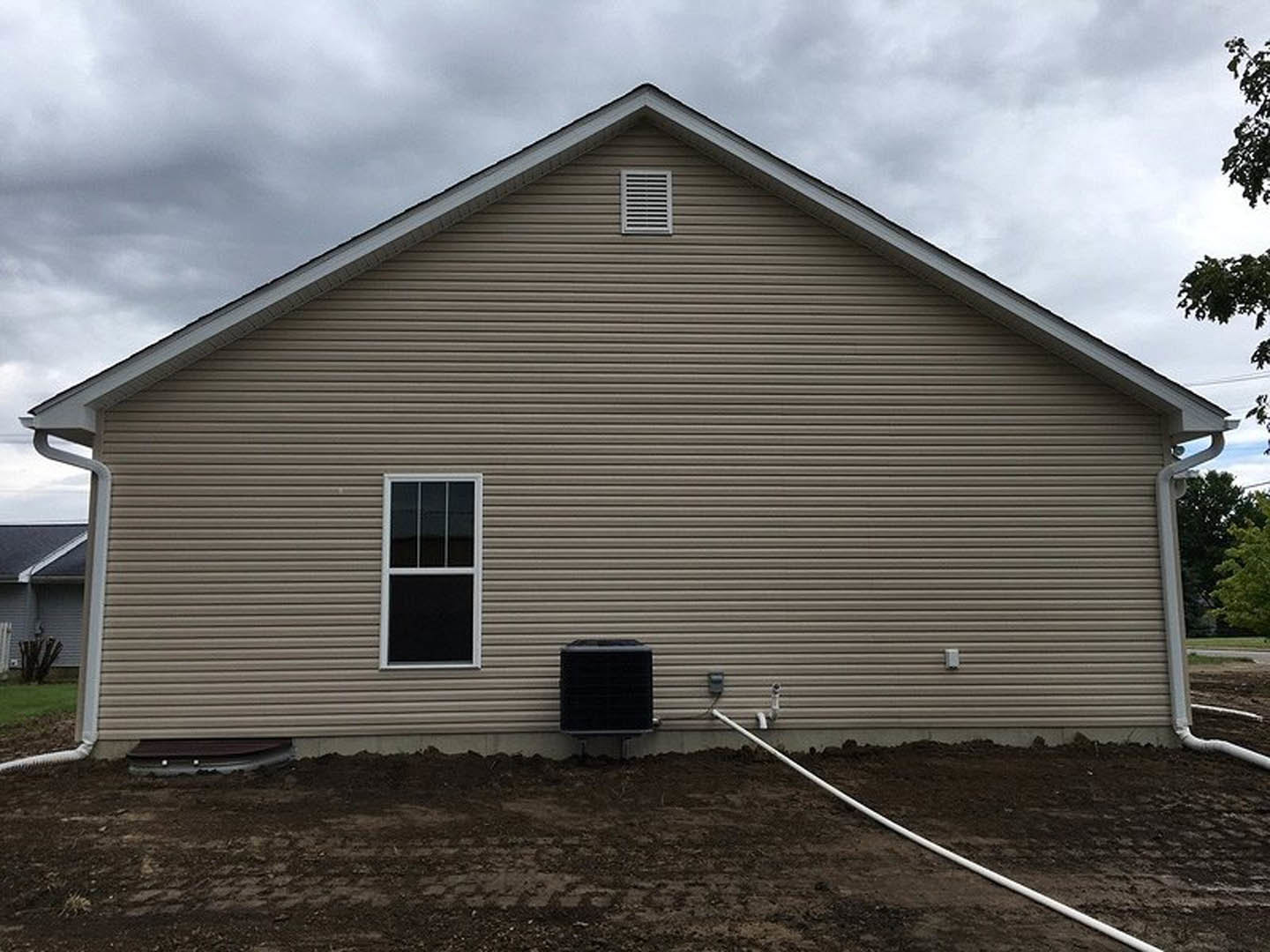 Light gray siding with a white-framed window, wall vent, black hose reel mounted beside a metal pole, and a dirt patch along the foundation.