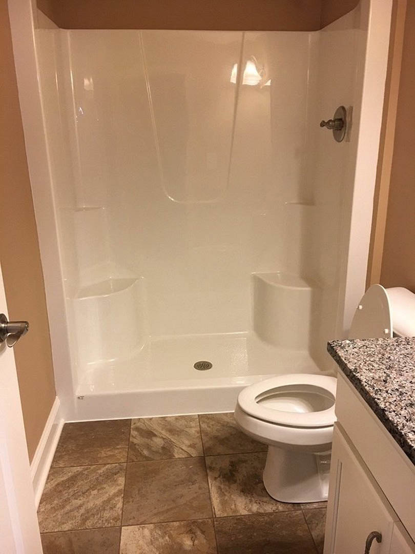 White toilet and walk-in shower with glass door, brown tile flooring, light-colored countertop with chrome faucet, neutral tiled walls