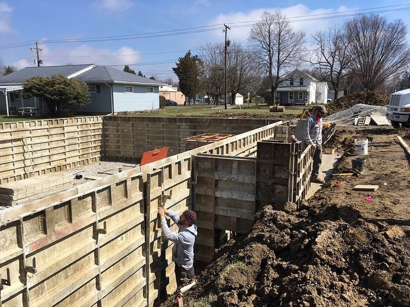 Men building a house foundation outdoors, with construction materials, a parked truck, and green trees in the background