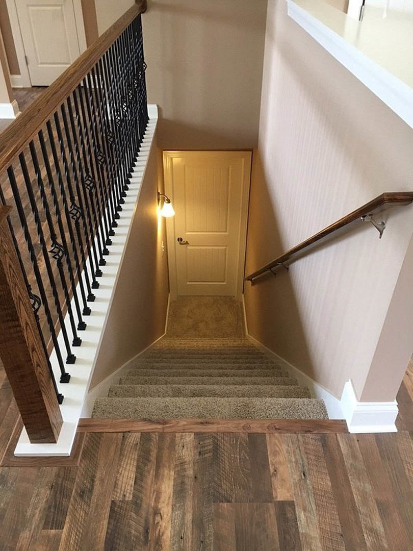 Hardwood staircase with white wall-mounted handrail, wood balusters, and a paneled door illuminated by a wall sconce; close-up of natural wood flooring