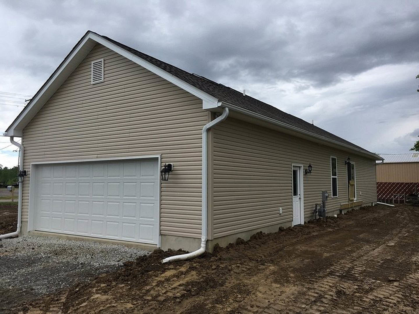 White siding house with attached garage, paneled garage door, rain gutter along shingled roof, small vent above entry, cloudy sky overhead.