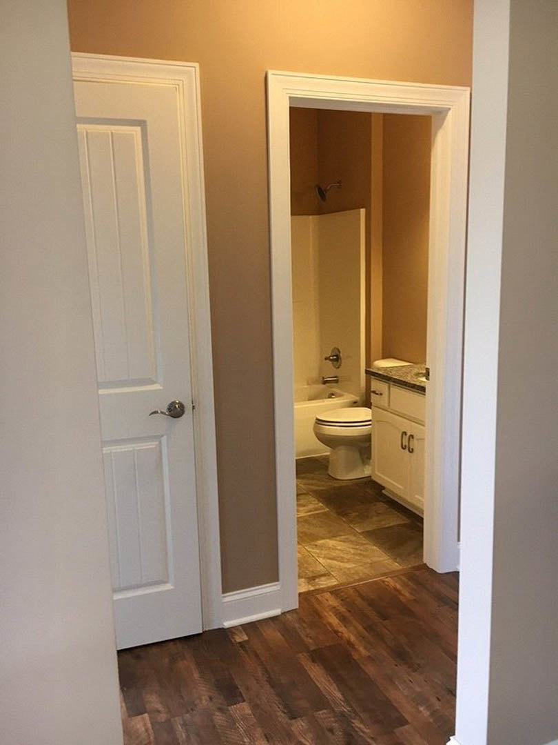 White porcelain toilet and rectangular sink with chrome faucet set against light gray tile walls, wood plank flooring, and a frameless mirror above the vanity