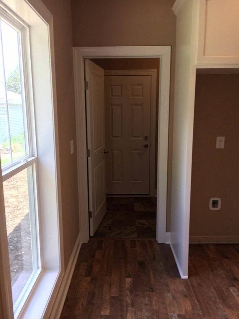 Hallway with light wood flooring, white walls, white door with silver handle, window on side wall showing outdoor view, white electrical outlet near baseboard