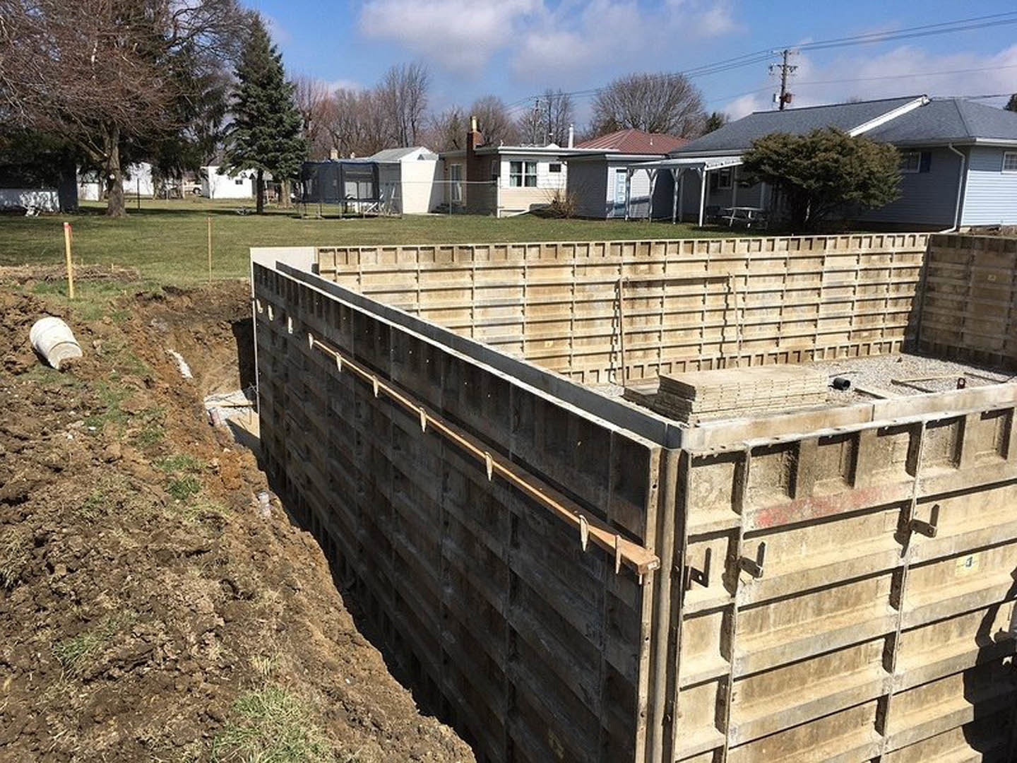 Concrete foundation under construction with exposed lumber framing, white bucket on dirt, green-leaved tree nearby, and house partially visible in background.
