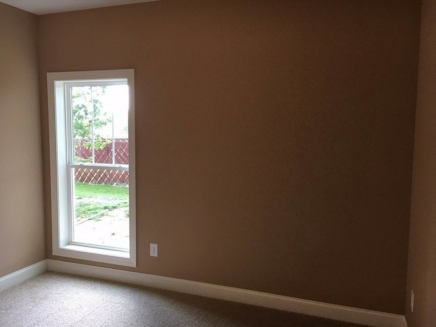 Carpeted room with white plaster walls, large window overlooking fenced yard with grass and trees