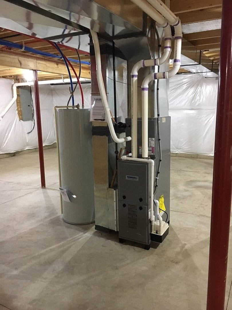 Large steel boiler with connected pipes in unfinished basement, exposed beams and concrete floor visible.