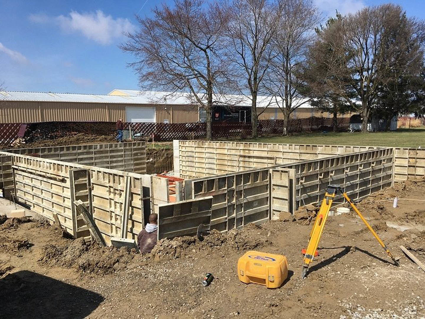 Framed white exterior wall with window, construction equipment and yellow plastic box on dirt foundation, worker kneeling near tree and fence under open sky