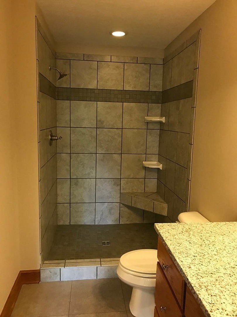 Modern bathroom featuring a glass-enclosed shower, white toilet beside a marbled countertop, brown cabinetry, and tiled flooring with a raised tile step