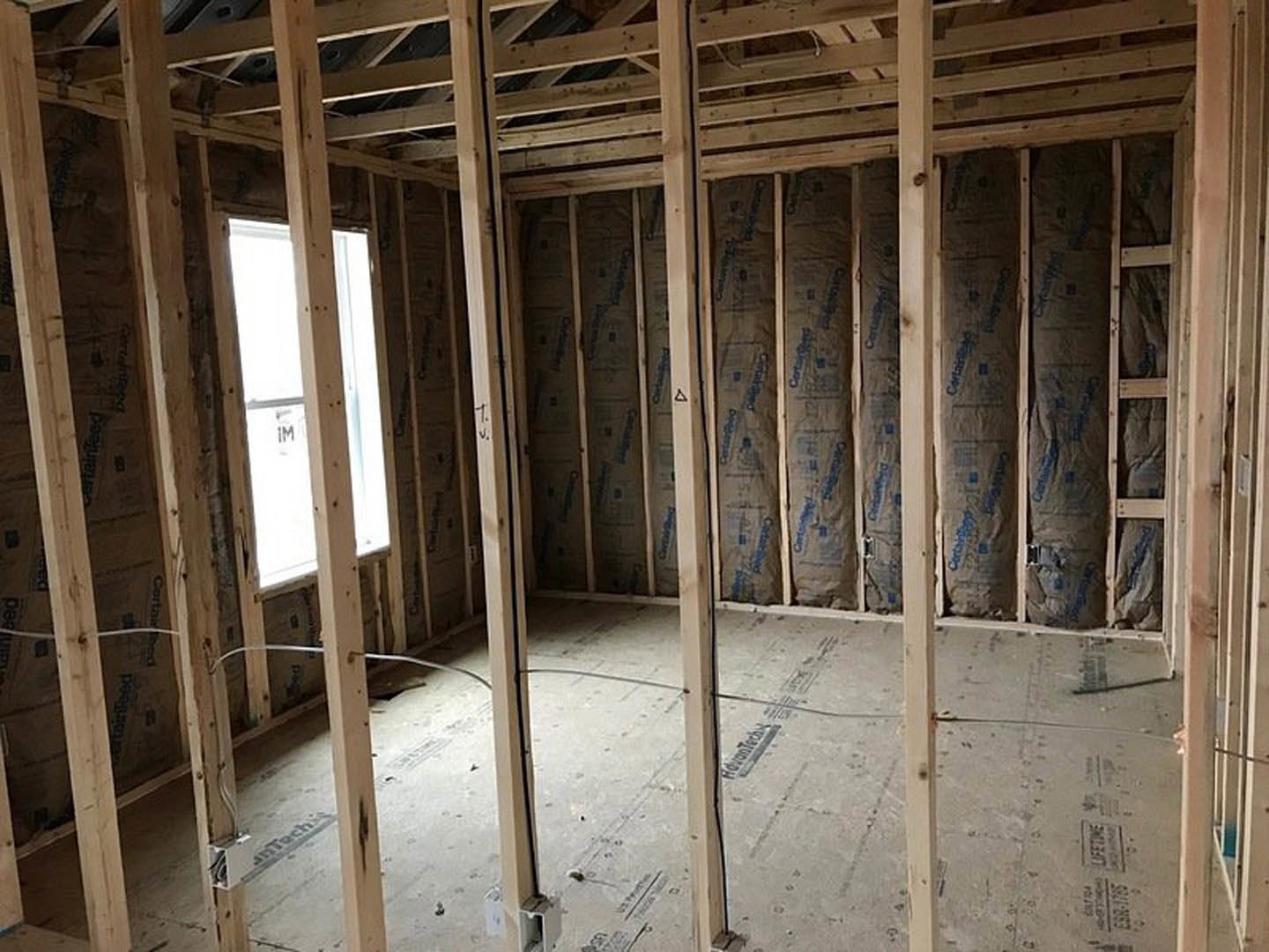 Wood-framed room under construction with exposed beams, window frame displaying a sign, wall lined with insulation bags, and wooden slats visible on the ceiling
