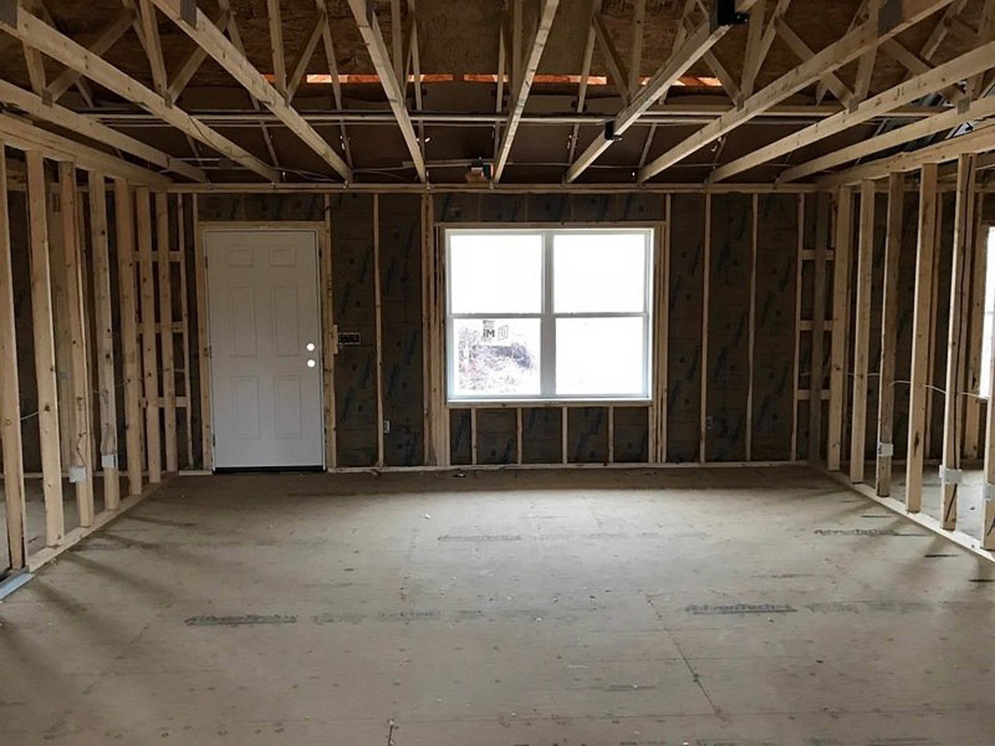 Room with concrete floor, exposed metal beams, white plaster ceiling, large window, and white door; natural daylight highlights wood framing and building insulation.