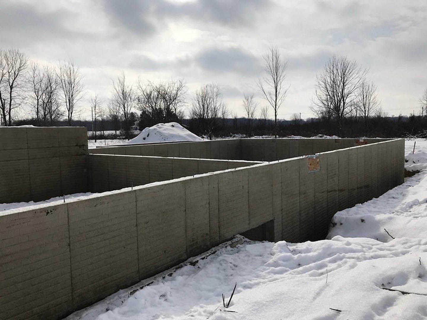 Concrete retaining wall with central opening, snow-covered ground, leafless trees in background, winter sky, brown square object with white frame near fence