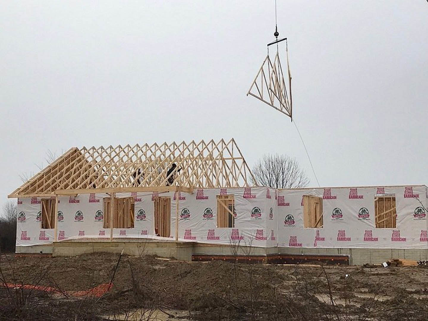 Wood-framed house under construction with exposed beams, white protective sheeting, crane overhead, piles of dirt and sticks on ground, metal structure visible in foreground