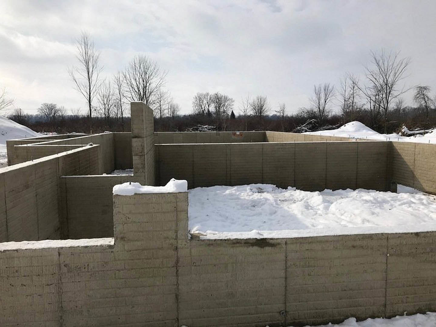 Wood-framed custom home under construction with snow covering the ground and partially blanketing exterior walls, leafless trees and cloudy sky in background