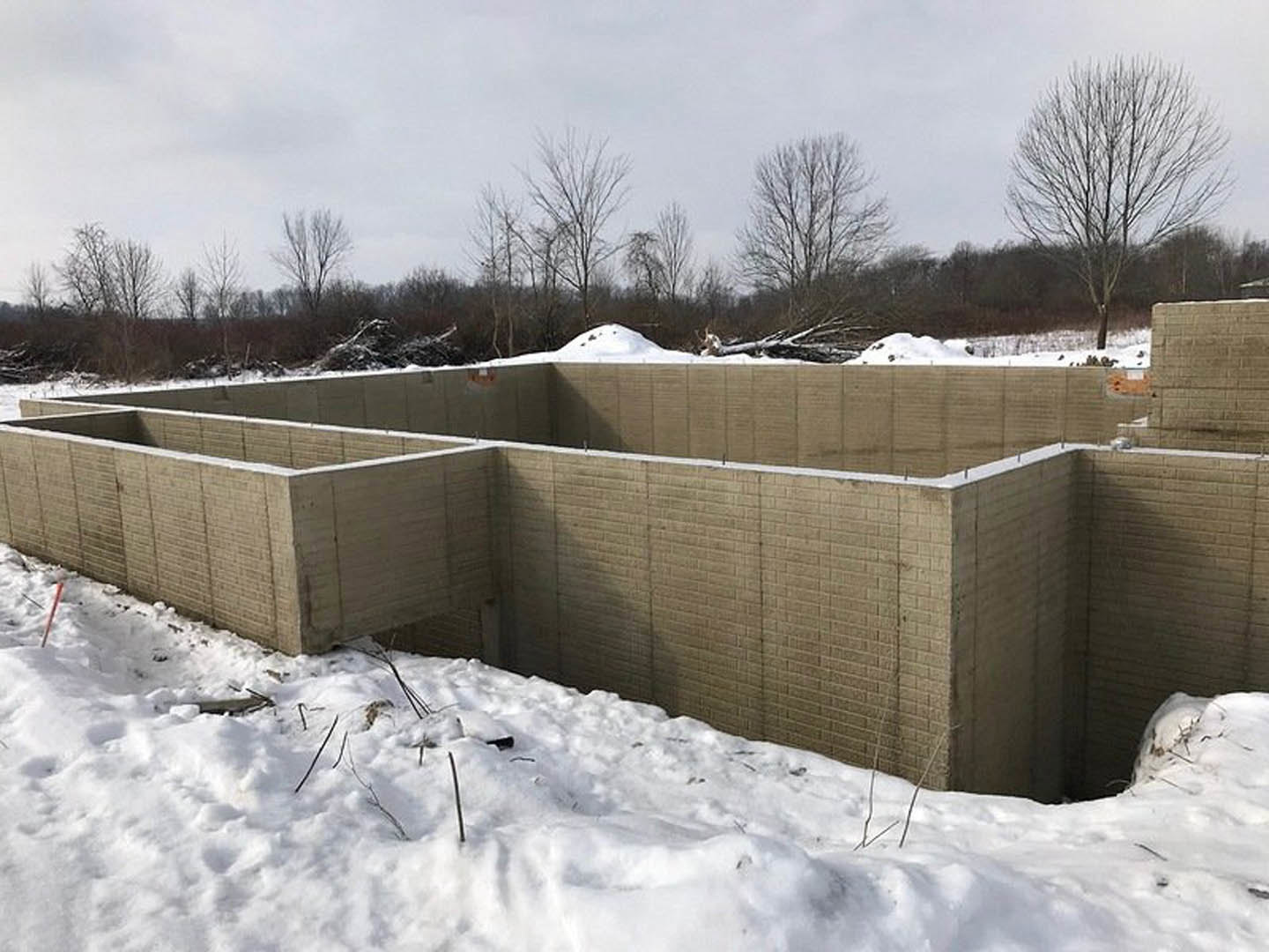 Concrete foundation and brick wall under construction on snow-covered ground, leafless trees and forest in background, overcast winter sky