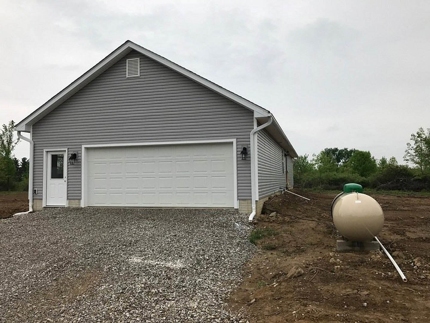 Brick house with white garage door, large round white propane tank with green top on front lawn, vent on exterior wall, white entry door with window, grassy yard, cloudy sky.