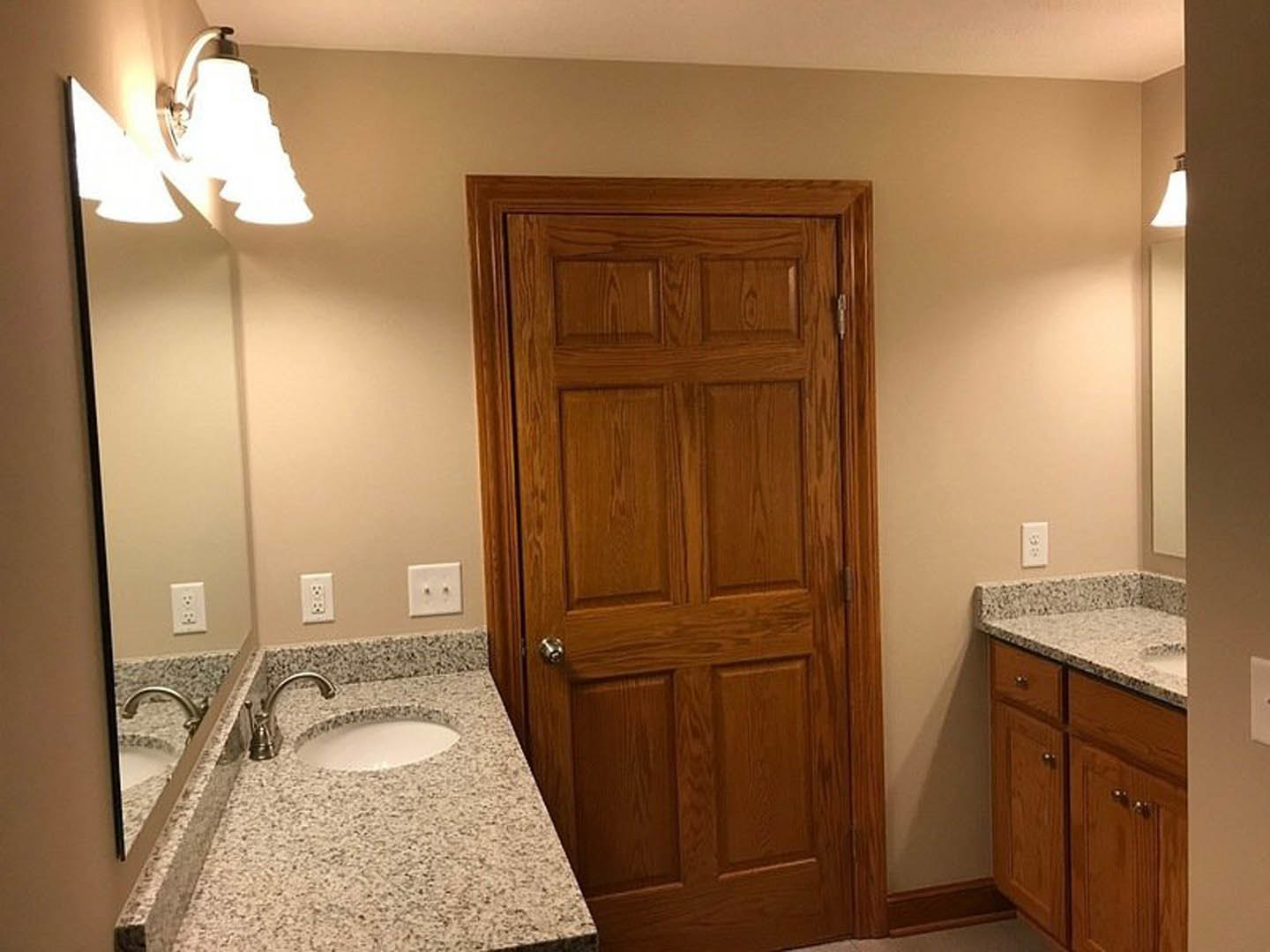 Bathroom with double vanity sinks, white countertops, wooden door with silver knob, tile flooring, wall outlet, modern faucets, and multi-bulb light fixture above mirrors