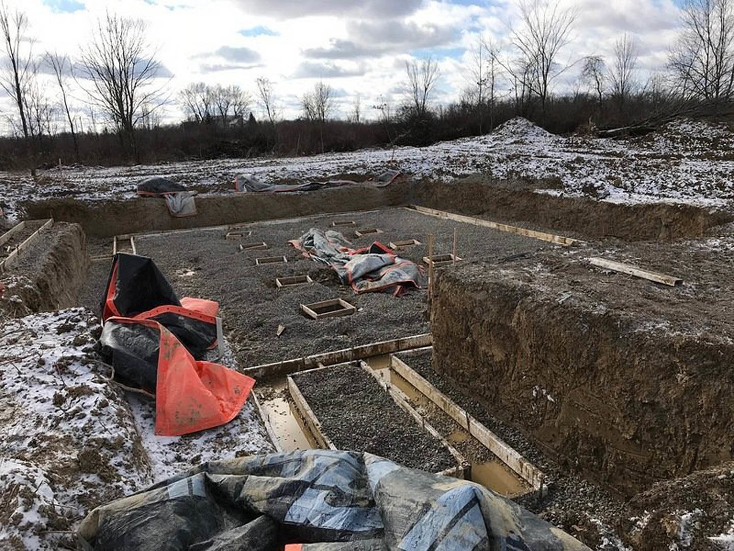 Construction site with large dirt pile, black and orange tarps covering ground, cloudy sky, surrounding trees, scattered gravel and plastic bag