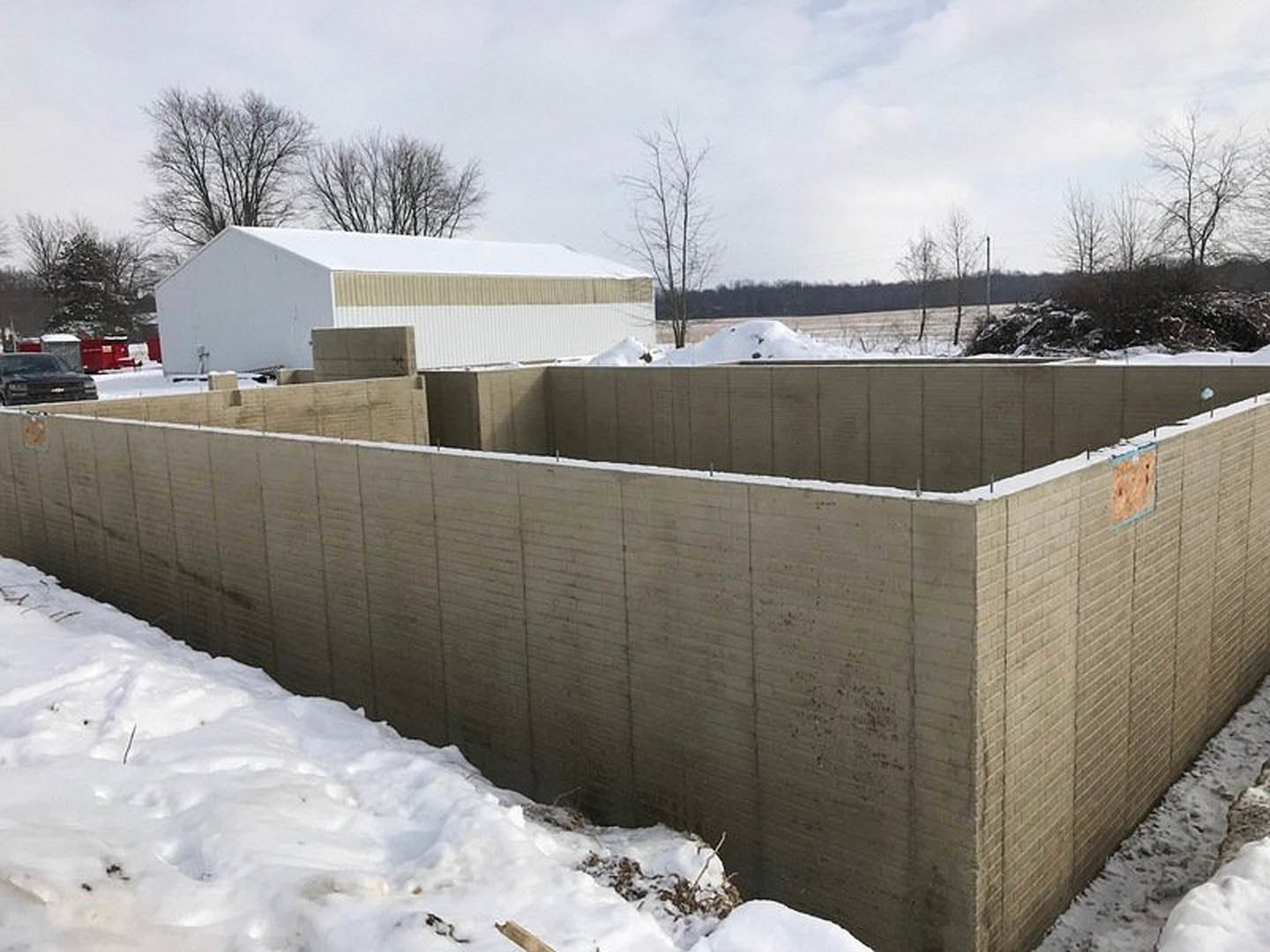 White custom home under construction with concrete walls, snow-covered roof, and surrounding trees in winter.