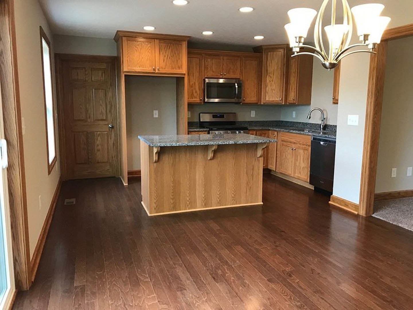 Open kitchen with wide-plank wood flooring, large central island with wood countertop, white cabinetry, stainless steel microwave, modern chandelier, and paneled door.