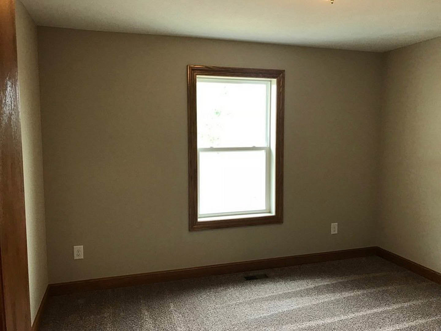 Bright room with white plaster walls, square window letting in natural light, light-colored carpet flooring, and wood trim details