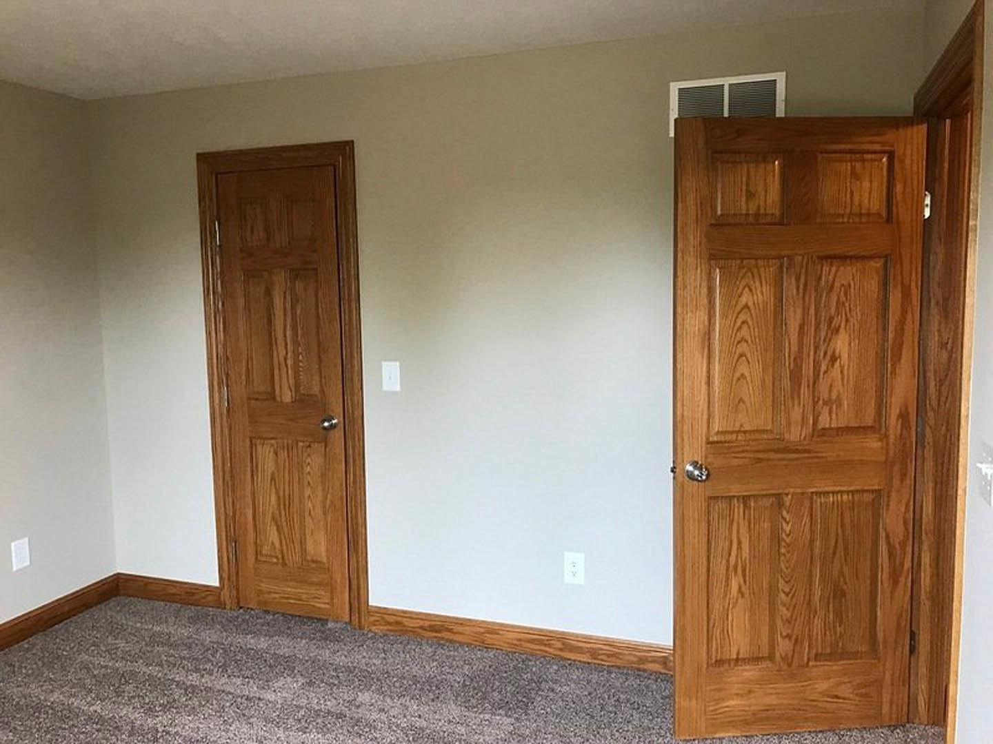 White-walled room with two wooden doors featuring silver handles, beige carpet flooring, wall vent, and close-up of wood grain surface