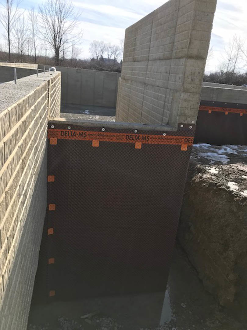 Large concrete exterior wall with orange tape and black sign, surrounded by bare trees and winter ground under a cloudy sky